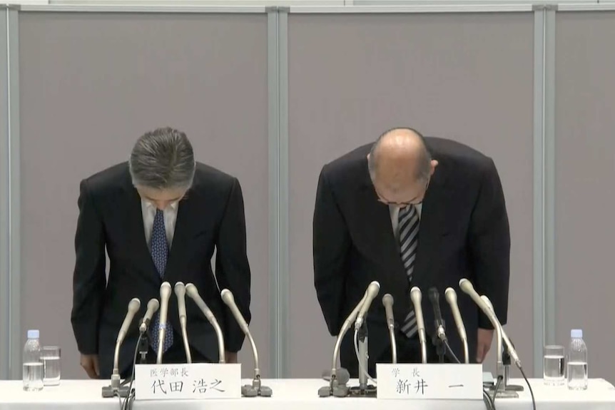 Two greying men in suits bow while stood behind a table during a press conference in Japan, with a grey wall behind them.