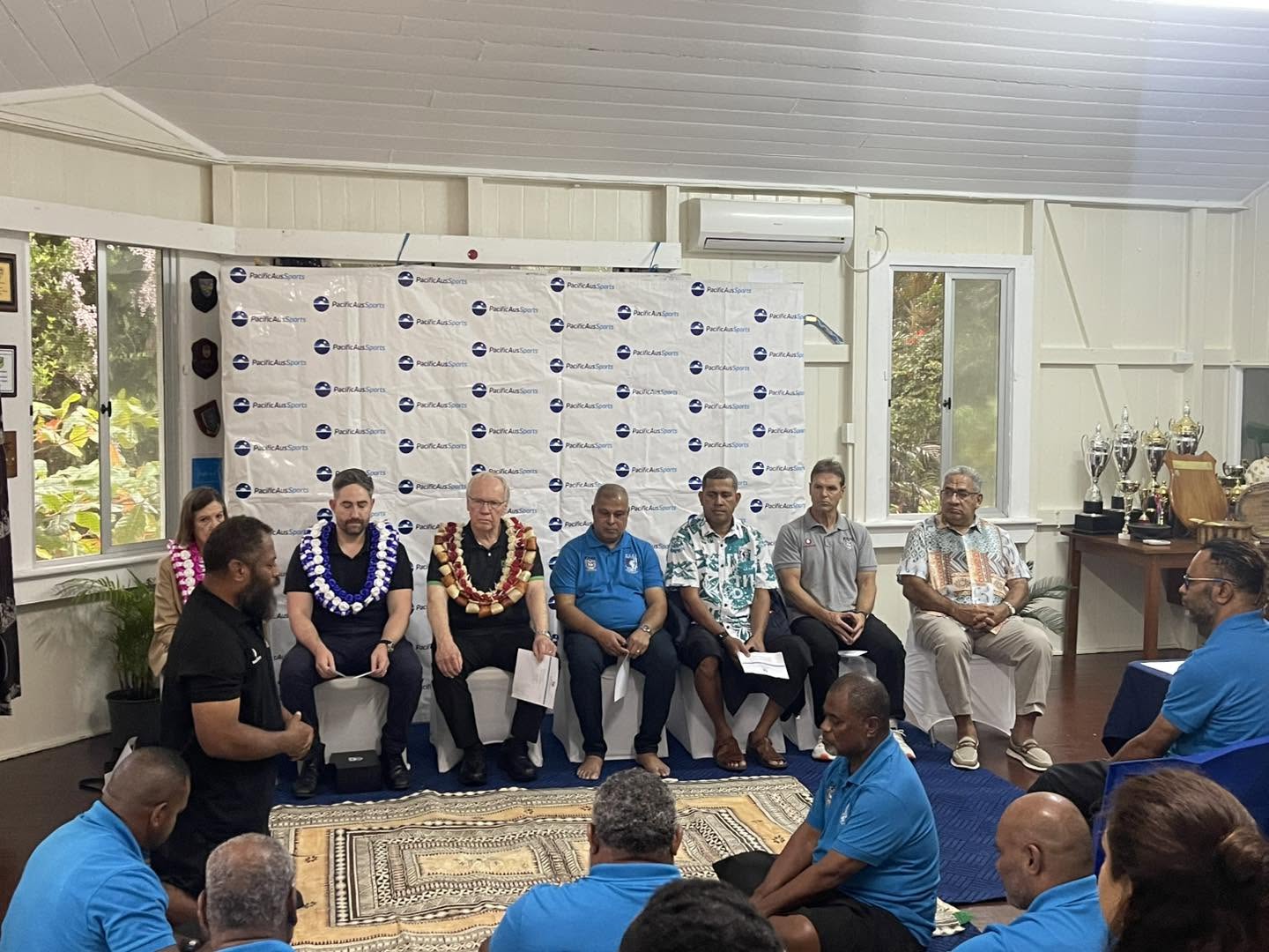 A Fijian man kneels on masi/tapa laid out on the floor in front of a line of men sitting on chairs in a room in Fiji with others