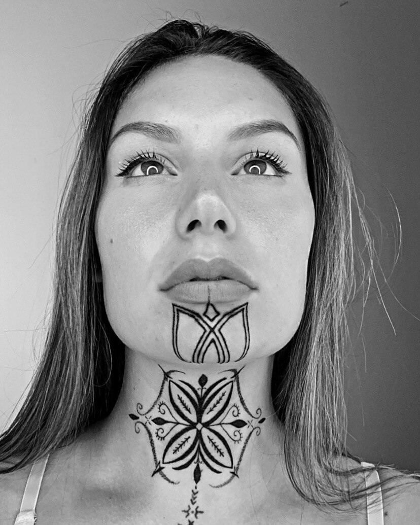 This black and white image shows a woman looking upwards. She has tattoos on her chin and neck.