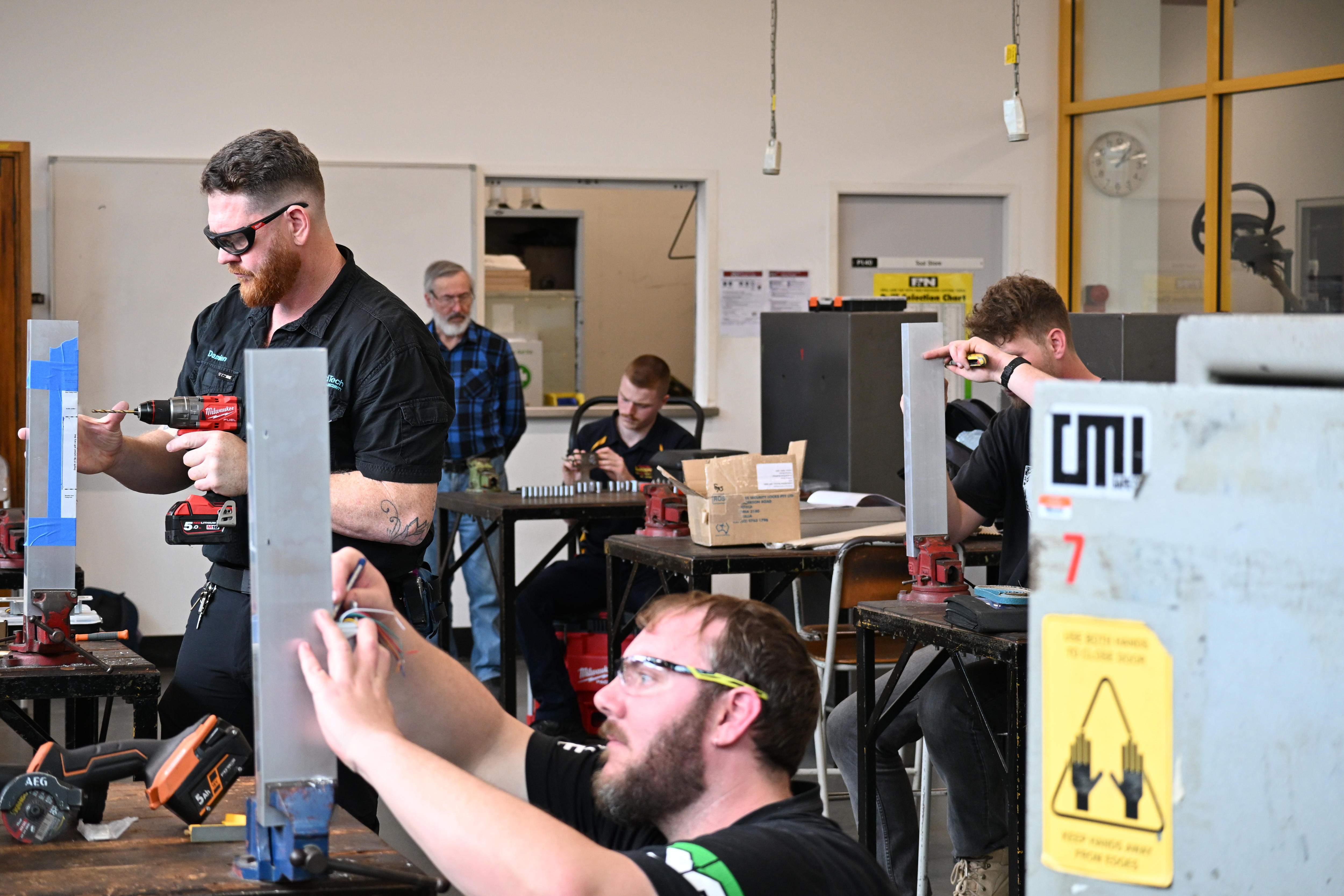 Four male locksmith apprentices use drills and tools in a workshop.