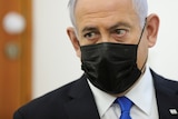 An elderly man with white hair in a black face mask and suit looks seriously to the side.