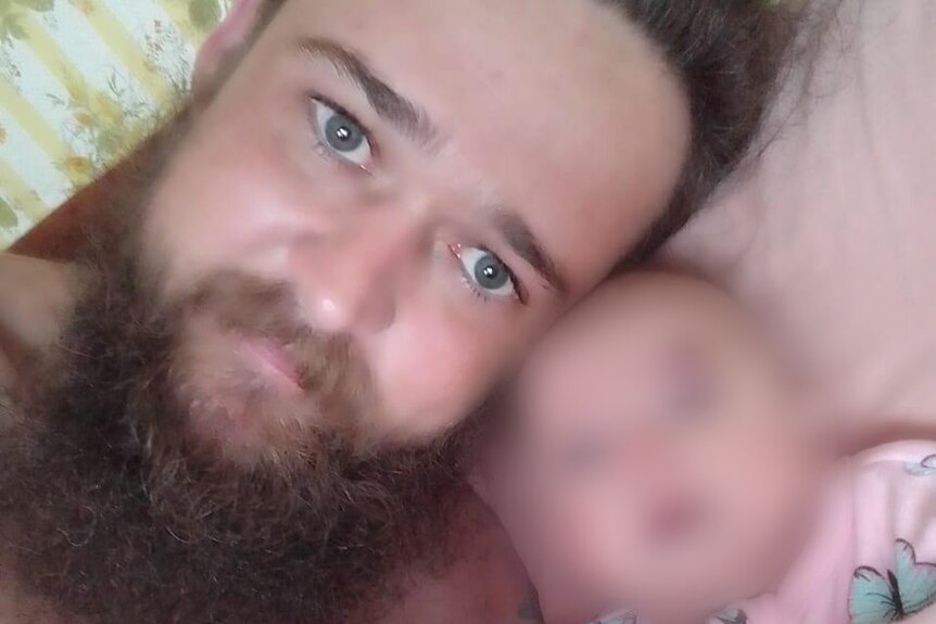 A man with a beard and moustache and a baby lying next to him with face blurred