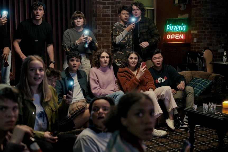A group of Gen Z people sit on couches around a seance table, holding phones up to record the action.