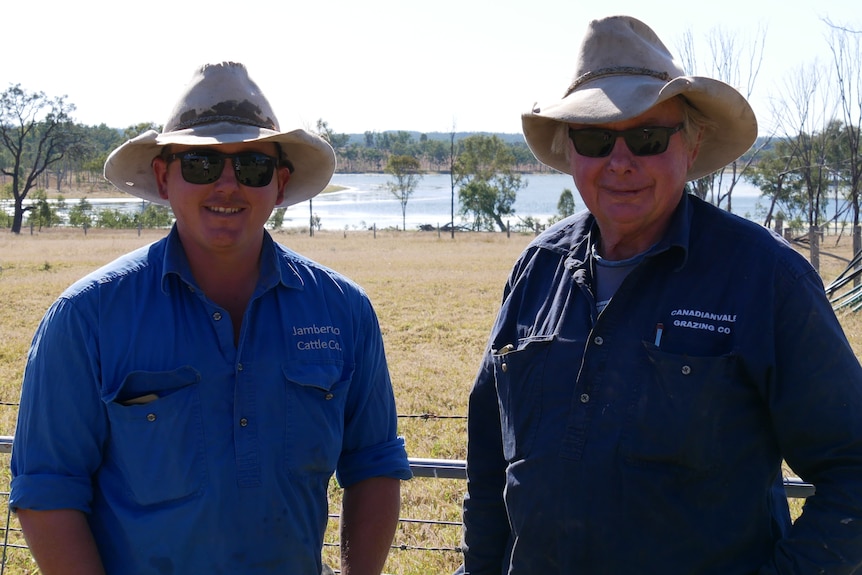 Two men both in blue work shirts stand smiling in front of a farm gate.