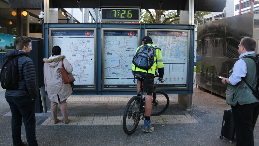 Two pedestrians and a man on a bike read large printed maps and timetables of Canberra's new bus network.
