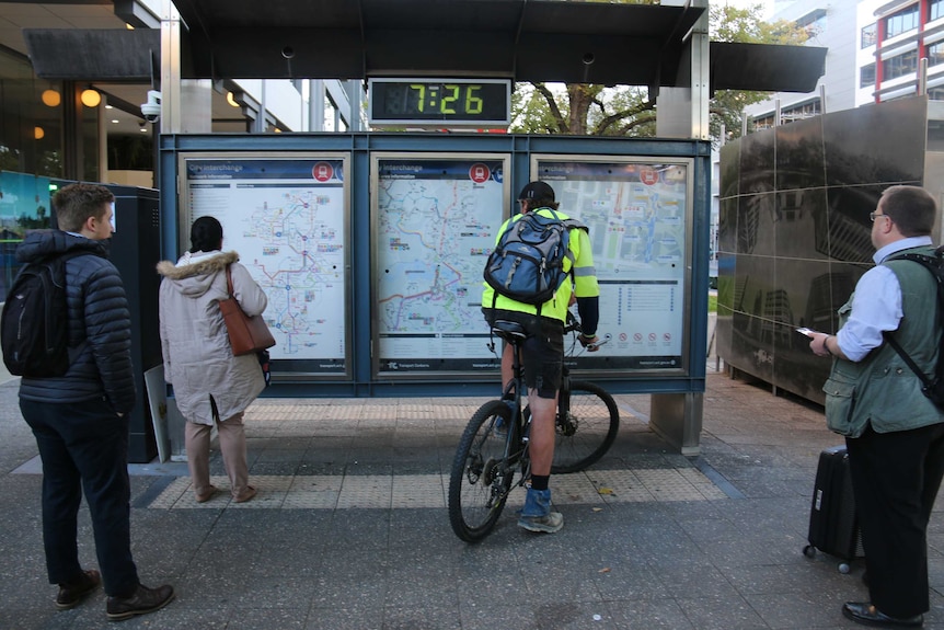 Two pedestrians and a man on a bike read large printed maps and timetables of Canberra's new bus network.