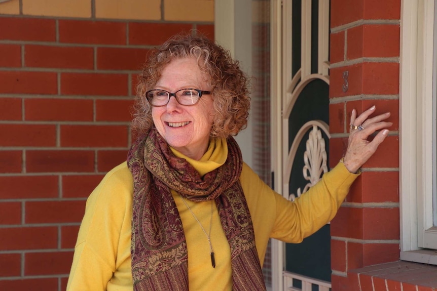 A woman wearing glasses and a patterned maroon scarf smiles while standing before the front door of a brick house.