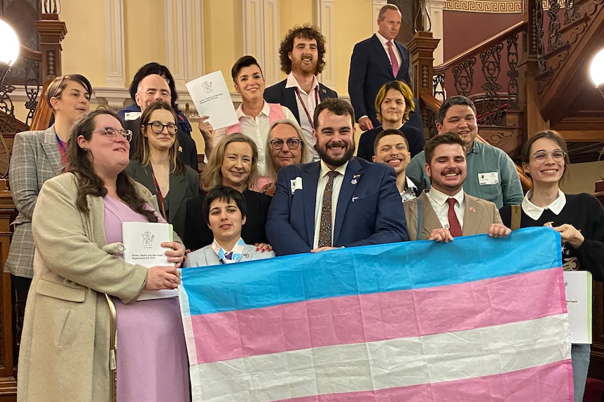 a group of people smiling on the steps of parliament and holding a trans flag