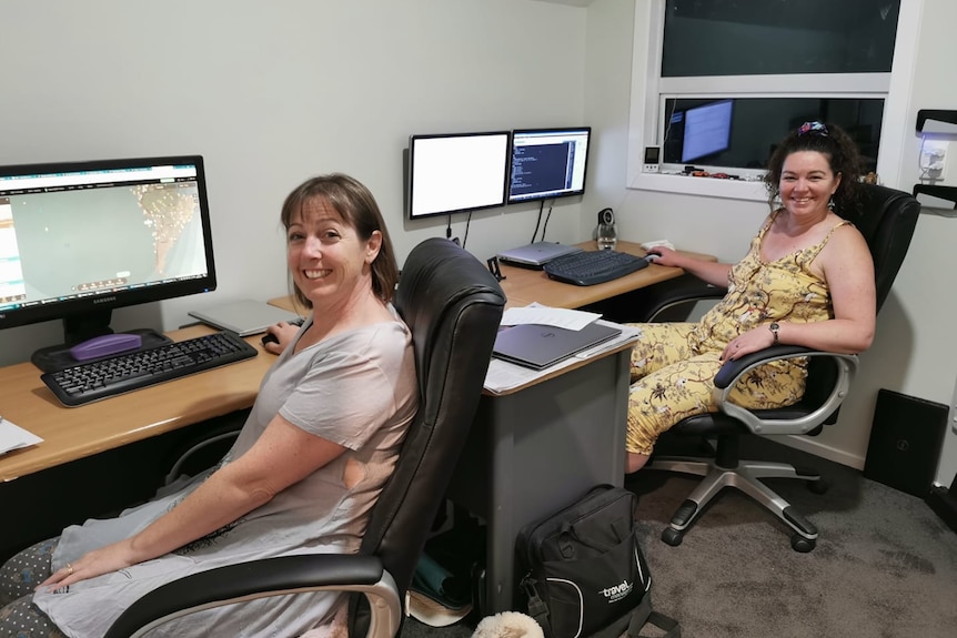 Two casually dressed, smiling middle-aged women sit at computer desks in a room at night.