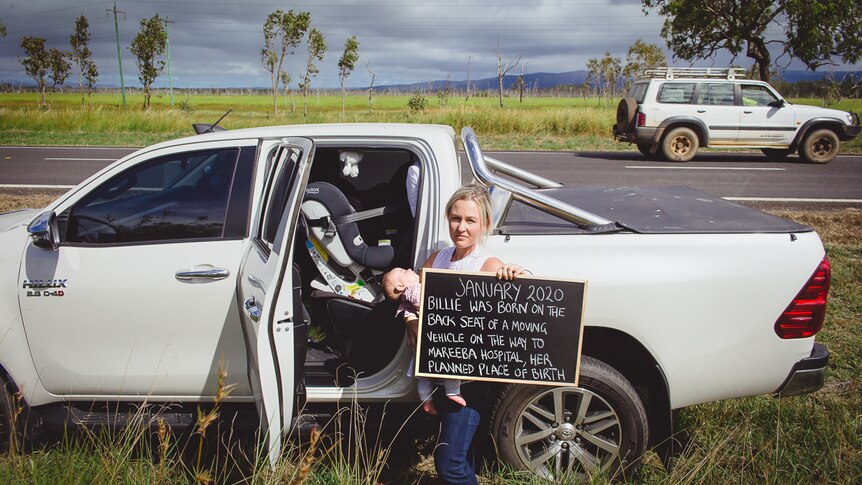Woman holding baby and sign standing in front of ute on regional highway