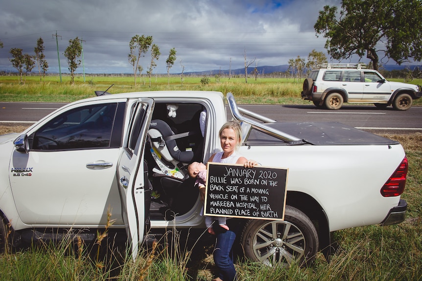 Woman holding baby and sign standing in front of ute on regional highway