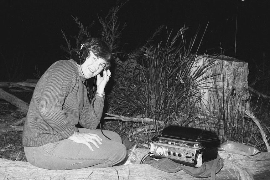 A woman who is Patsy Littlejohn wears headphones connected to a recorder and smiles as she sits on a log.