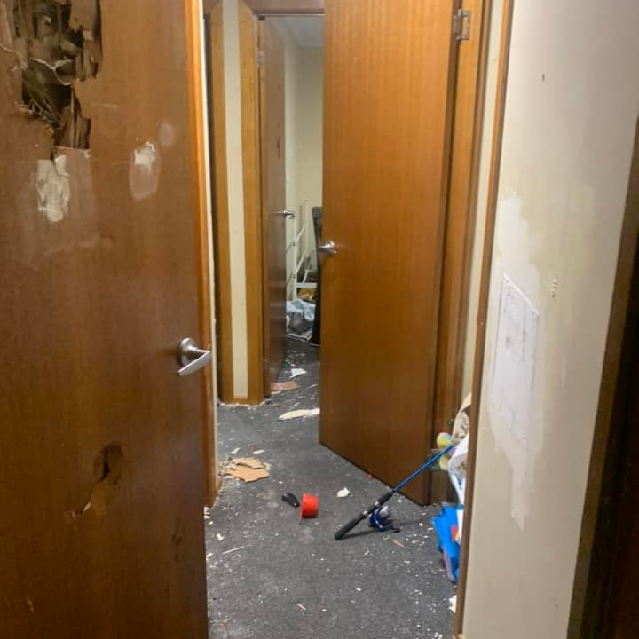 A hallway in a social housing unit trashed, walls broken, damaged doors hang open, floor messy