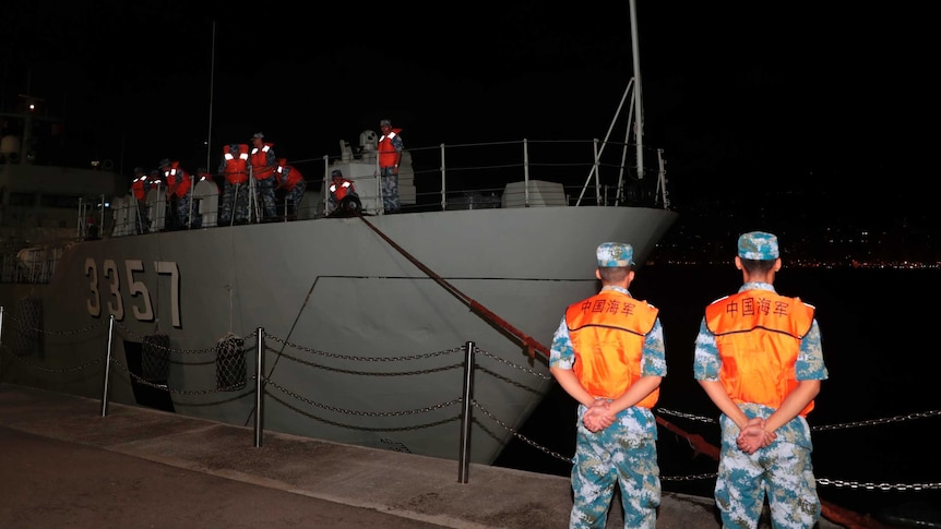 Two Chinese troops in camouflage and high-vis stand with their hands behind their backs as a boat carrying other troops docks.