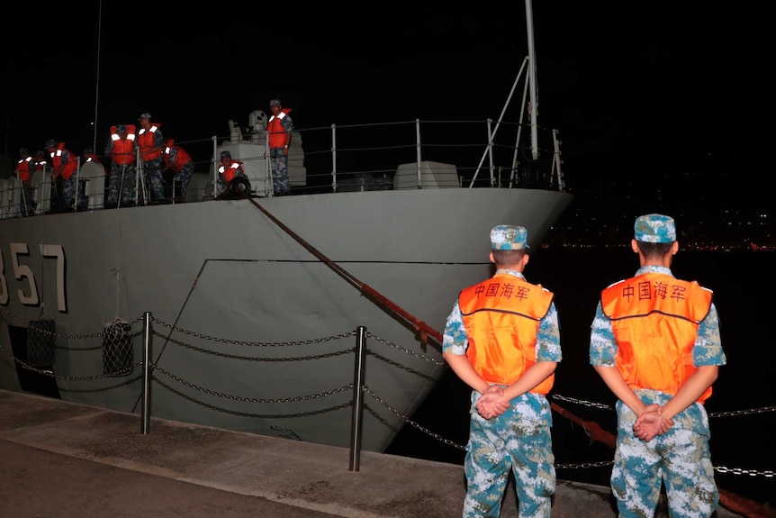 Two Chinese troops in camouflage and high-vis stand with their hands behind their backs as a boat carrying other troops docks.