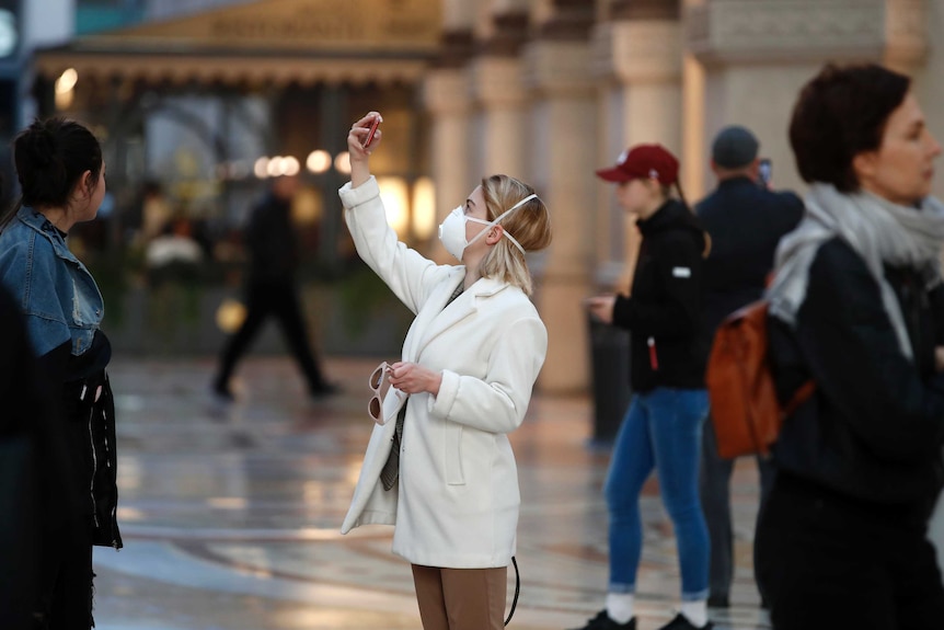A woman in a white jacket and wearing a sanitary mask holds up her phone.