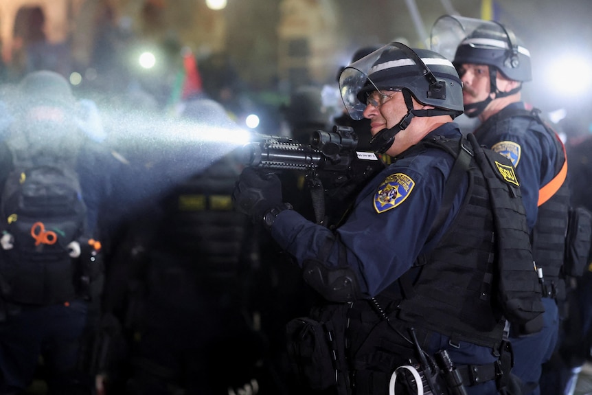 Ein Polizist richtet eine Waffe auf etwas, das außer Schussweite ist, während er vor einer Barrikade steht