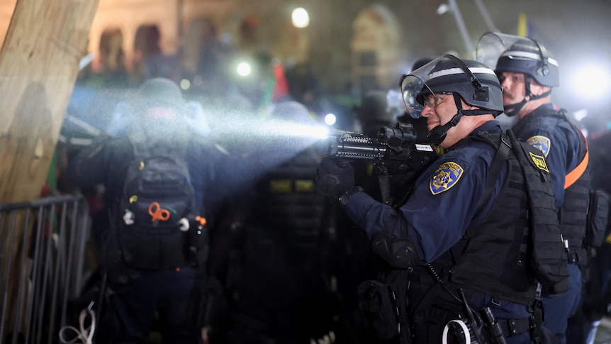 A policeman points a weapon toward something out of shot while standing in front of a barricade
