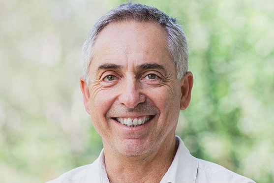 A man in his late fifties smiles into the camera wearing a white shirt and his hair in a short crop.