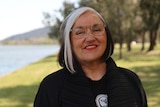 Margo Ngawa Neale, who has silver and brown hair, clear framed spectacles and red lipstick, poses for a photo by a body of water