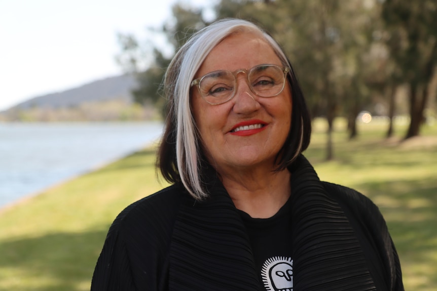 Margo Ngawa Neale, who has silver and brown hair, clear framed spectacles and red lipstick, poses for a photo by a body of water