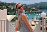 A woman wearing sunglasses smiles in front of the ocean with green hills in the distance