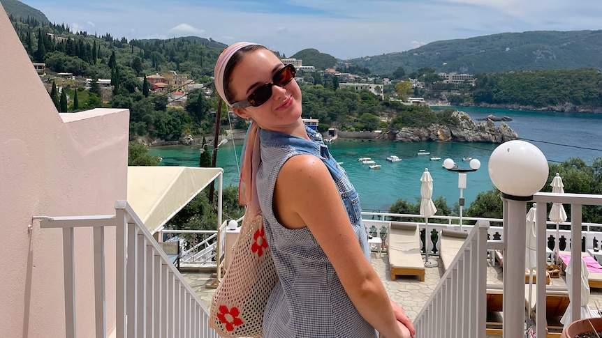 A woman wearing sunglasses smiles in front of the ocean with green hills in the distance