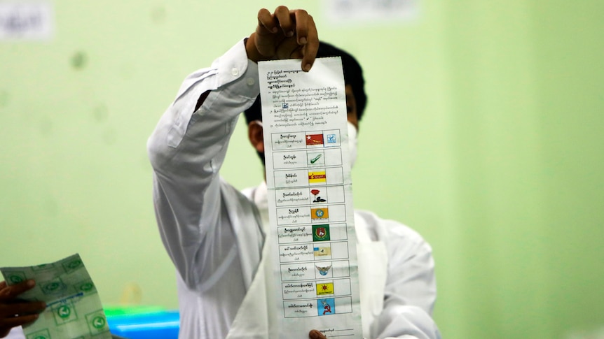 An official holds a long ballot paper aloft and counts the results makred in boxes next to coloured political party logos.