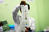 An official holds a long ballot paper aloft and counts the results makred in boxes next to coloured political party logos.