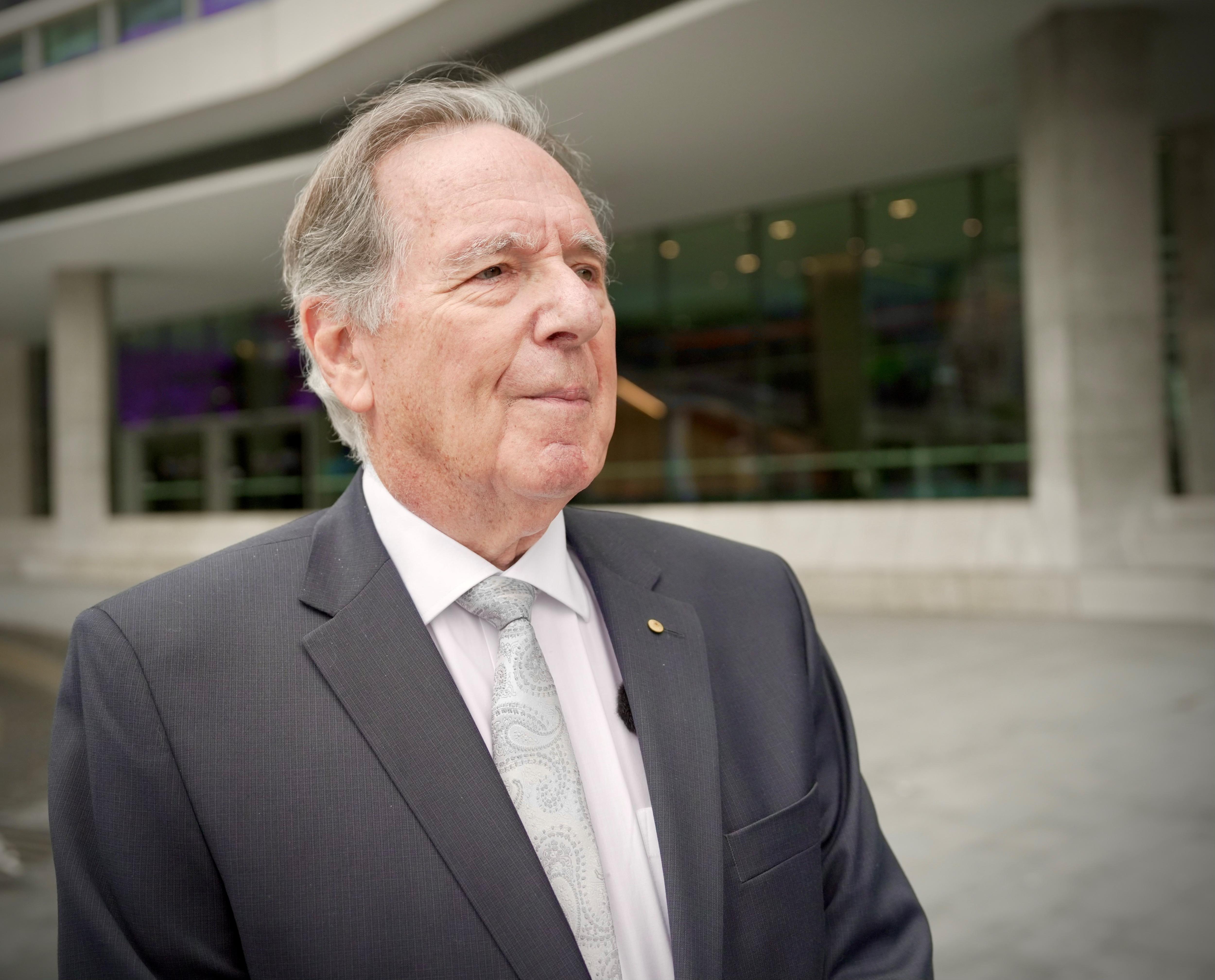 An older man wearing a suit and tie looks off into the distance in front of an unmarked building.