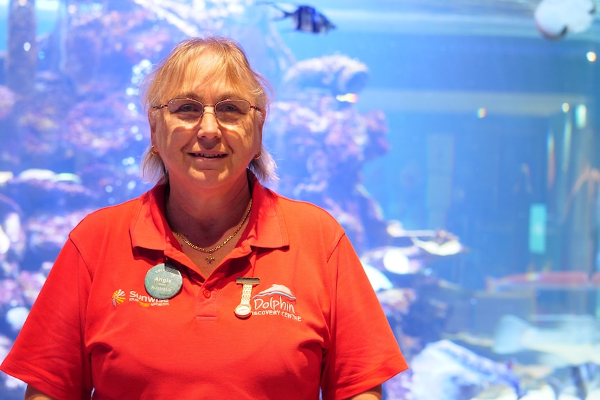 Older woman in bright red shirt in from of an aquarium, watch strapped to the tee, Bunburry Dolphin recovery centre lable.