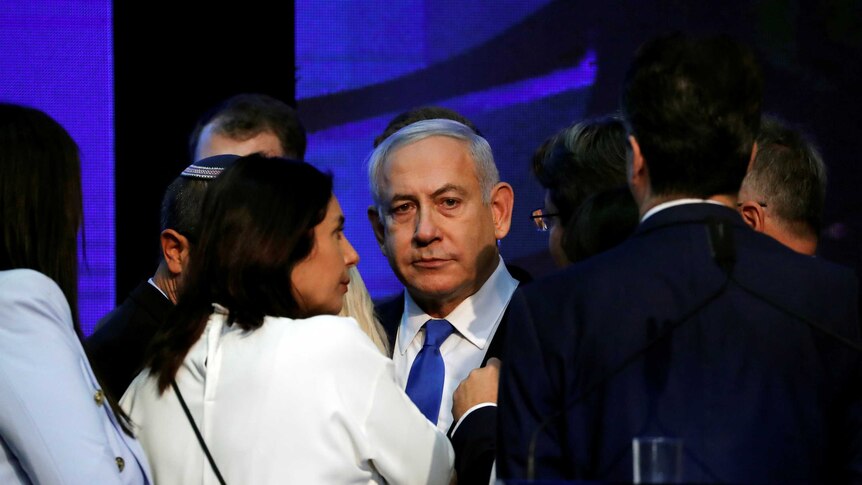 Israeli Prime Minister Benjamin Netanyahu looks past the camera in a crowd of people onstage in front of a blue background.