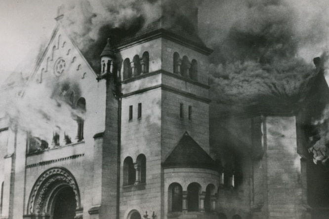 Black and white photo shows tall structure with arched windows and triangular roof with smoke emerging from all windows.