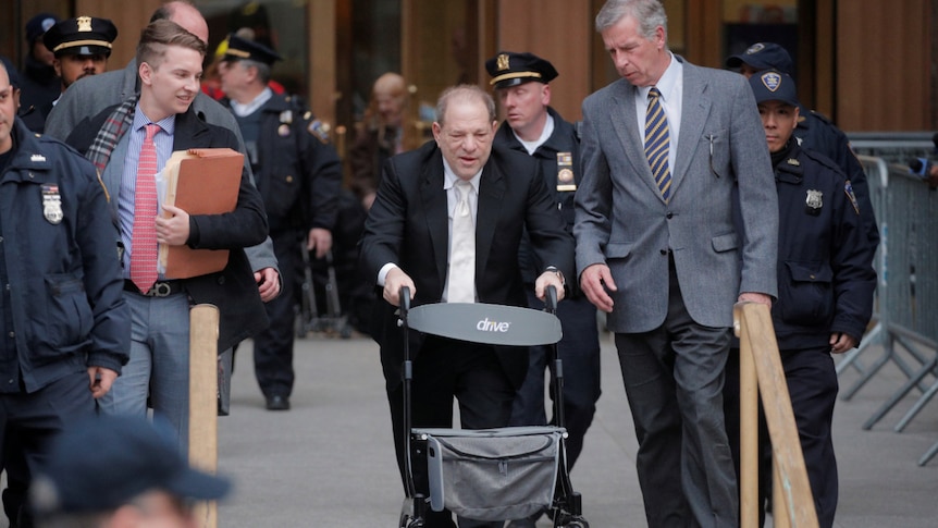 harvey weinstein walks in a suit with a trolley in front of him a man in a suit stands by his side and police officers behind