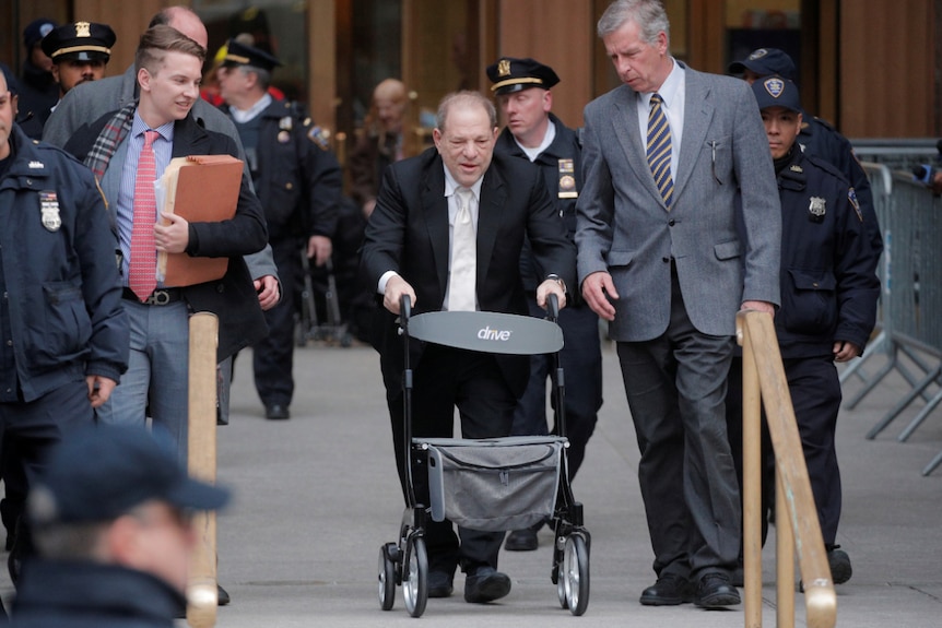 harvey weinstein walks in a suit with a trolley in front of him a man in a suit stands by his side and police officers behind