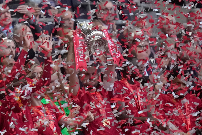 A grinning man dressed in red athletic garb holds a silver cup in the air filled with red and white strips of falling paper