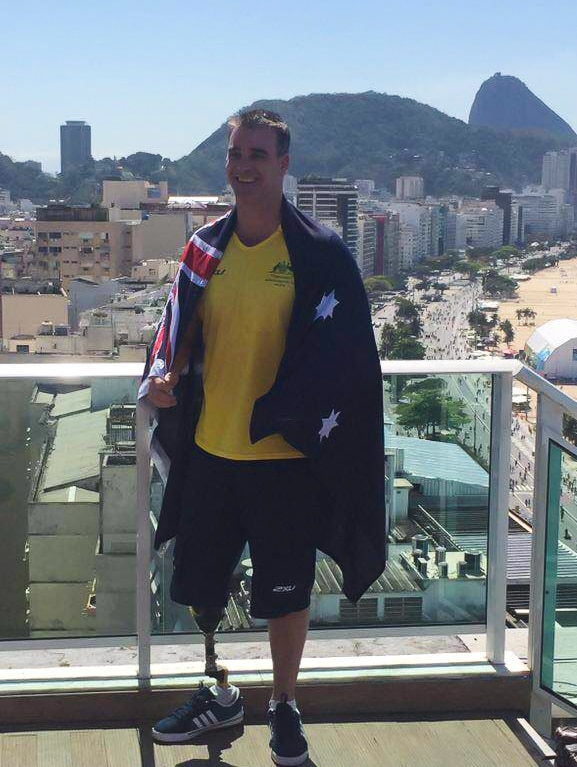 A man with a prosthetic right leg draped in the Australian flag in Rio with two men on the left