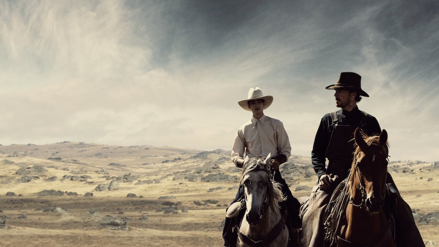 Two actors dressed as cowboys riding horses towards the camera, against a wilderness backdrop.
