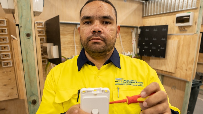A young indigenous man wears a high-vis shirt and holds a screwdriver and light socket in his hands
