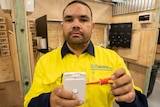 A young indigenous man wears a high-vis shirt and holds a screwdriver and light socket in his hands
