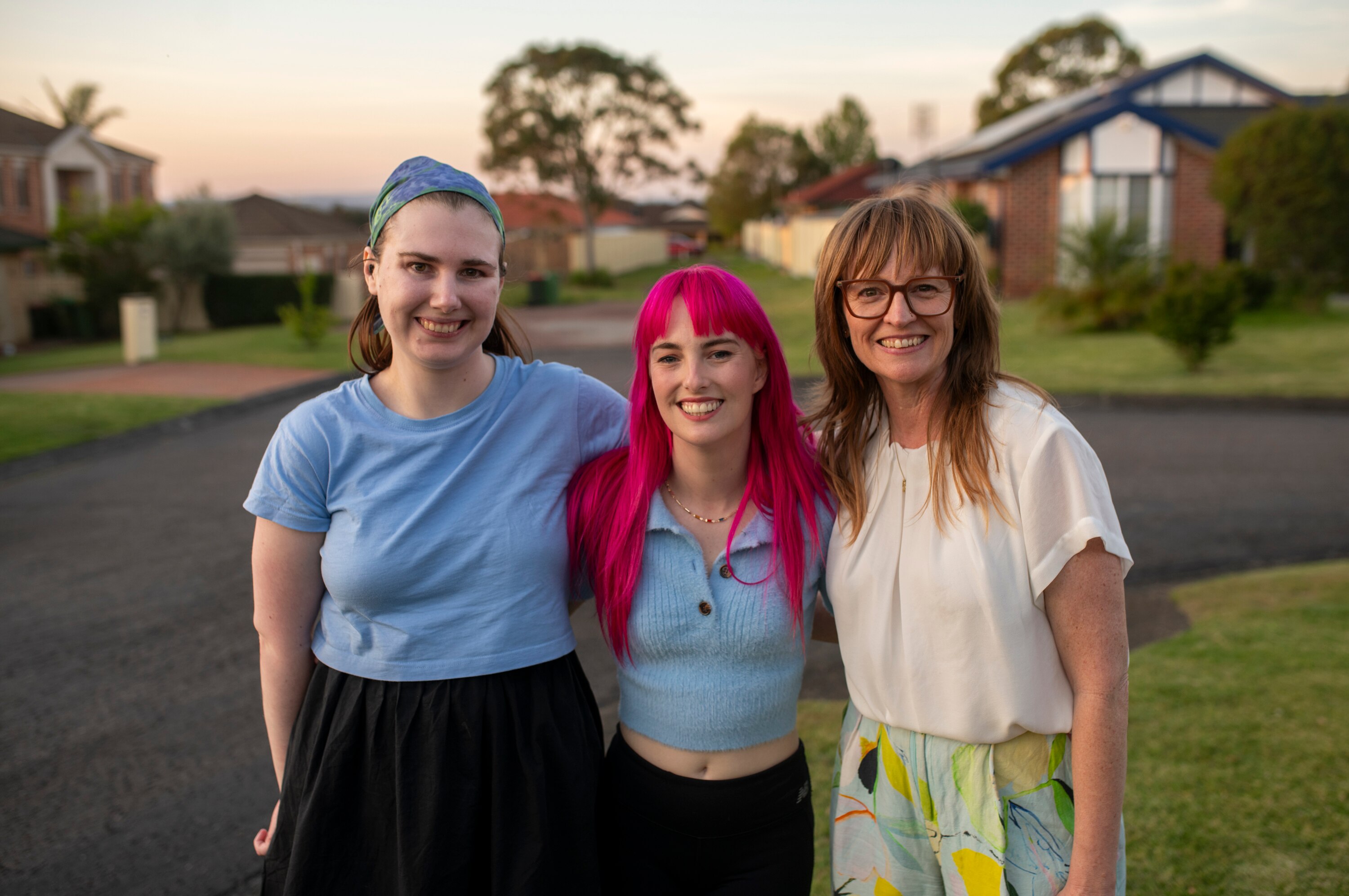 Três mulheres paradas em uma rua suburbana sorrindo. Um com blusa azul, outro com cabelo rosa e outro com óculos de leitura.