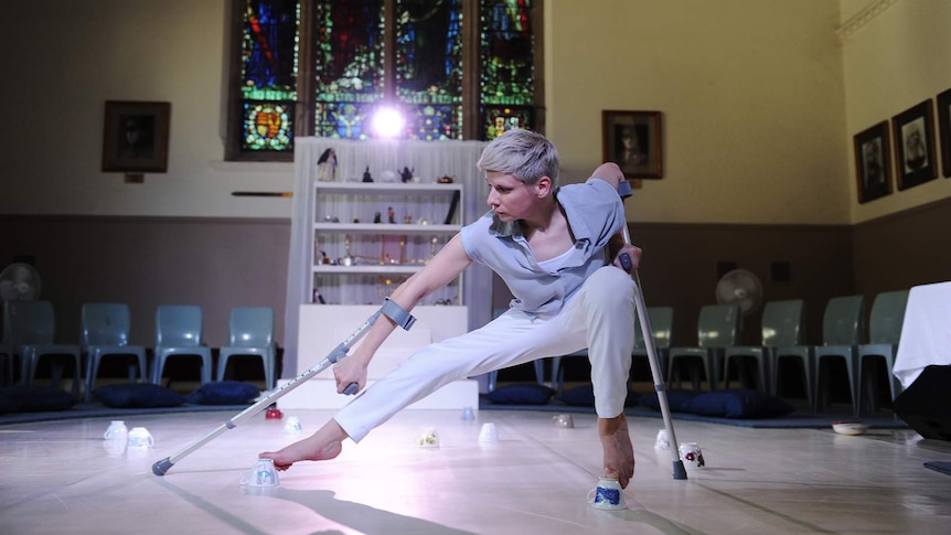 Woman with short blonde hair using crutches and dancing with her feet on upturned teacups, stained glass window behind.