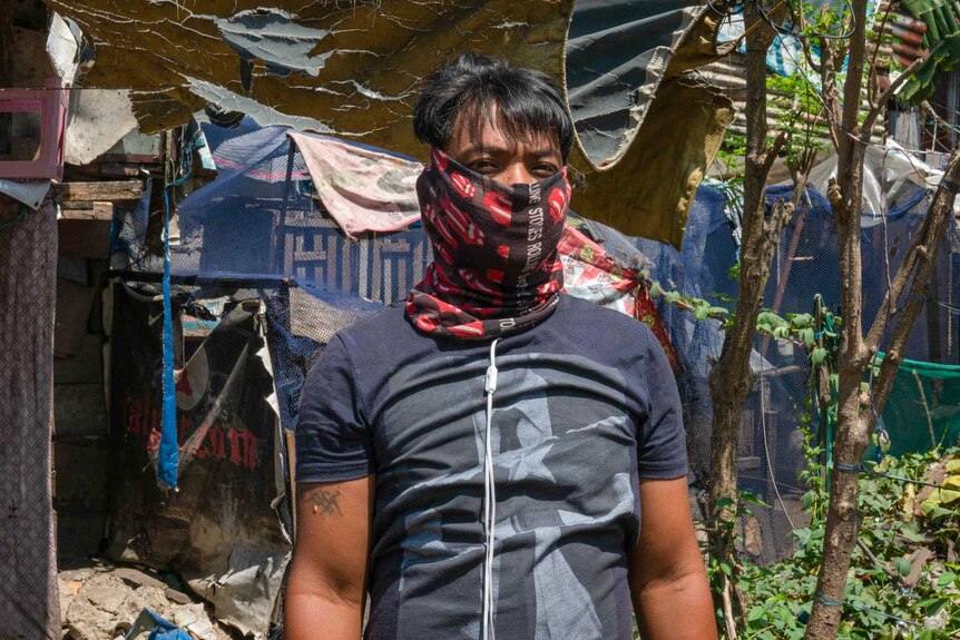 A man with a bandana wrapped around his face behind a decrepit row of houses