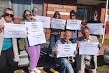 Six women and two men holding protest messages on large sheets of paper outside the red brick Tenterfield District Hospital