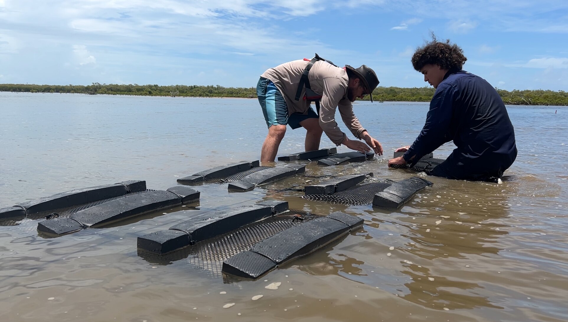 Two men crouch beside black foam pieces that are floating in ankle-deep water