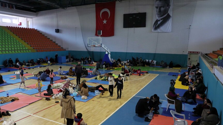 In a sports gymnasium, you view a group of people resting on its wooden basketball floor on brightly-coloured mats.