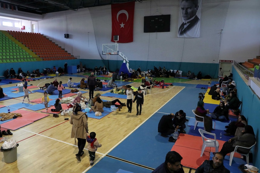 In a sports gymnasium, you view a group of people resting on its wooden basketball floor on brightly-coloured mats.