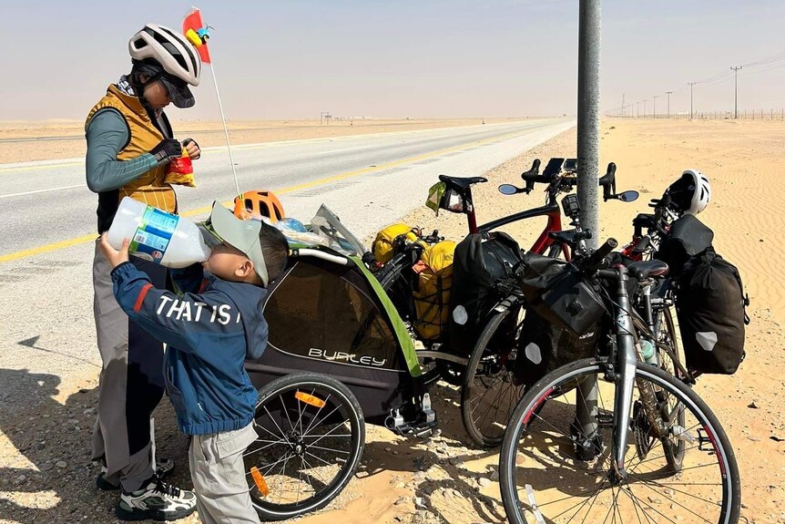 A woman and a young boy stop beside their bike next to a bitumen road through a sandy desert. 