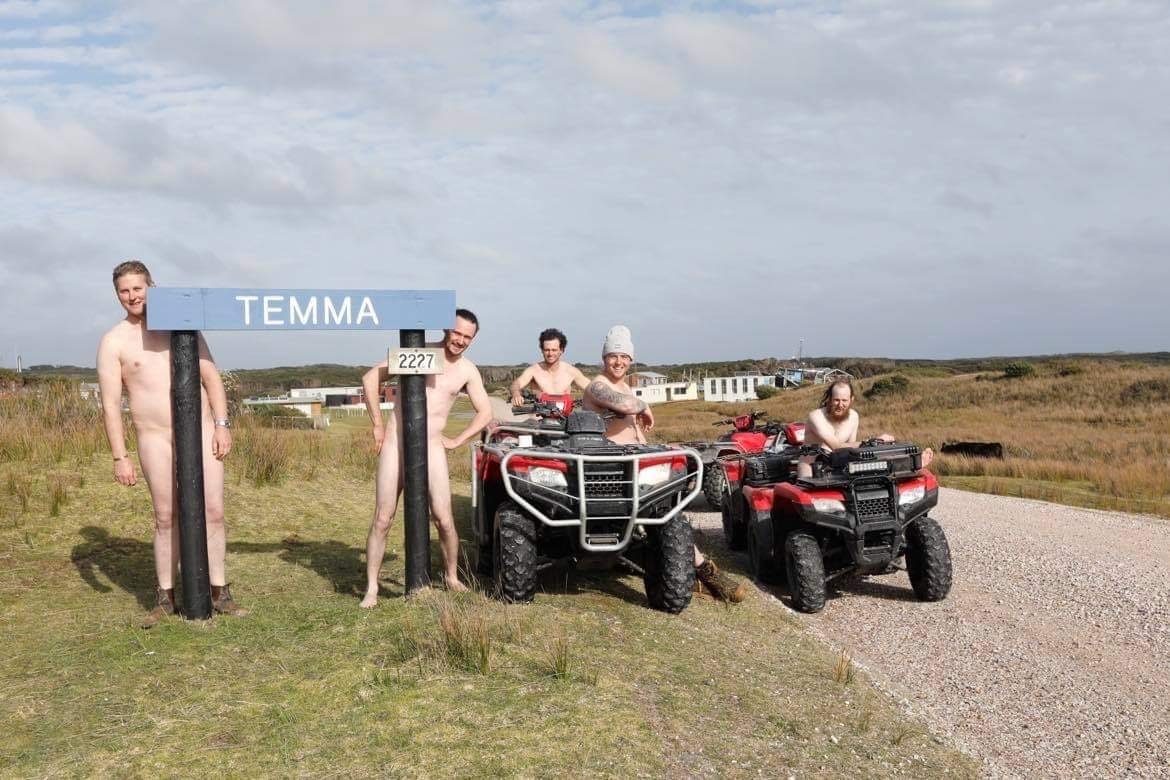 Five naked men standing or seated on four wheelers with small shack town in distance