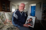 An old white man holding a photo of his dead wife. He is sitting on a couch